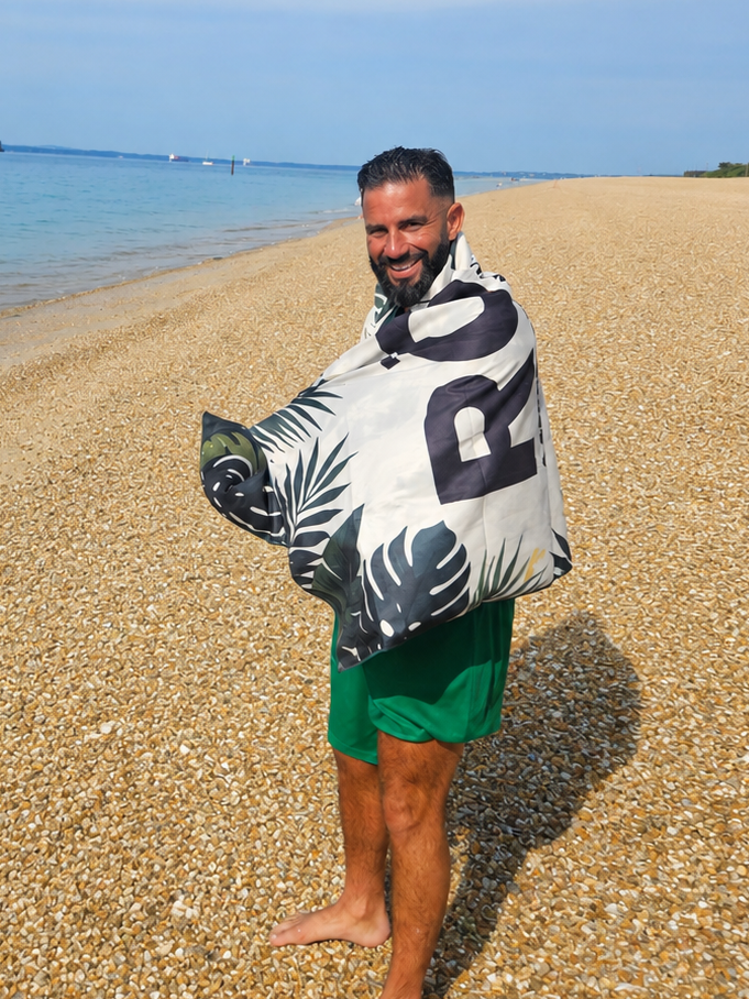 A man standing on a sandy beach wrapped in a white beach towel with a black tropical leaf pattern and large black numbers, wearing green swim shorts, with the blue sea and sky in the background.