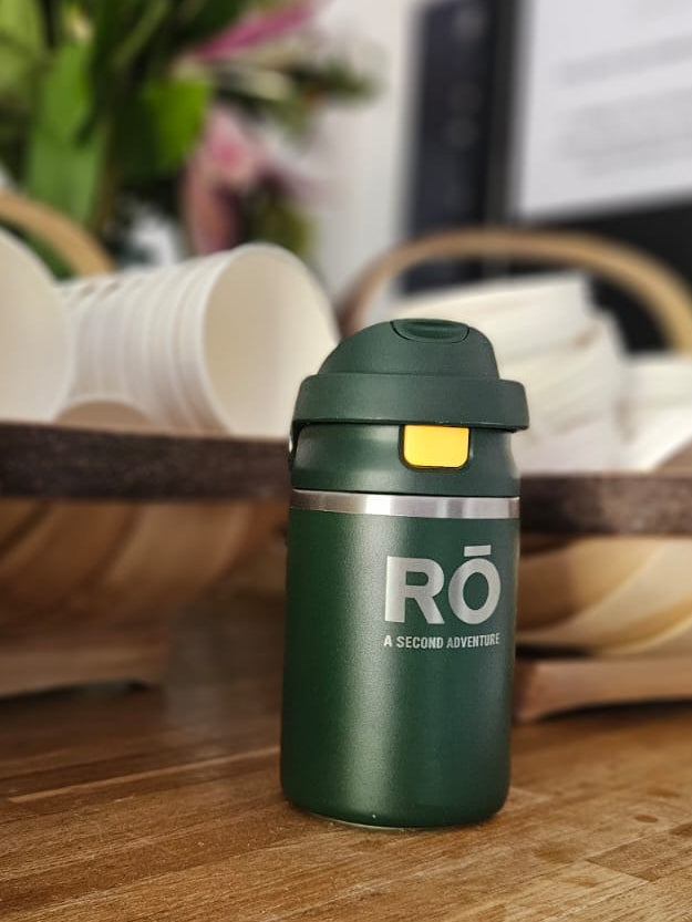 A dark green reusable insulated tumbler with a flip-top lid and a silver band reading 'RO A small carbon' sits on a wooden counter in a café or restaurant setting.