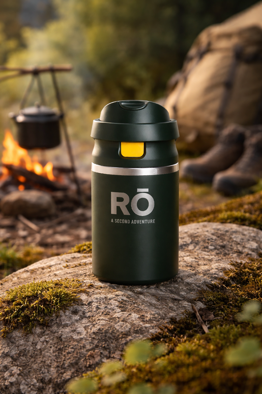 Black insulated bottle with yellow accent on a rock near a campfire and backpack