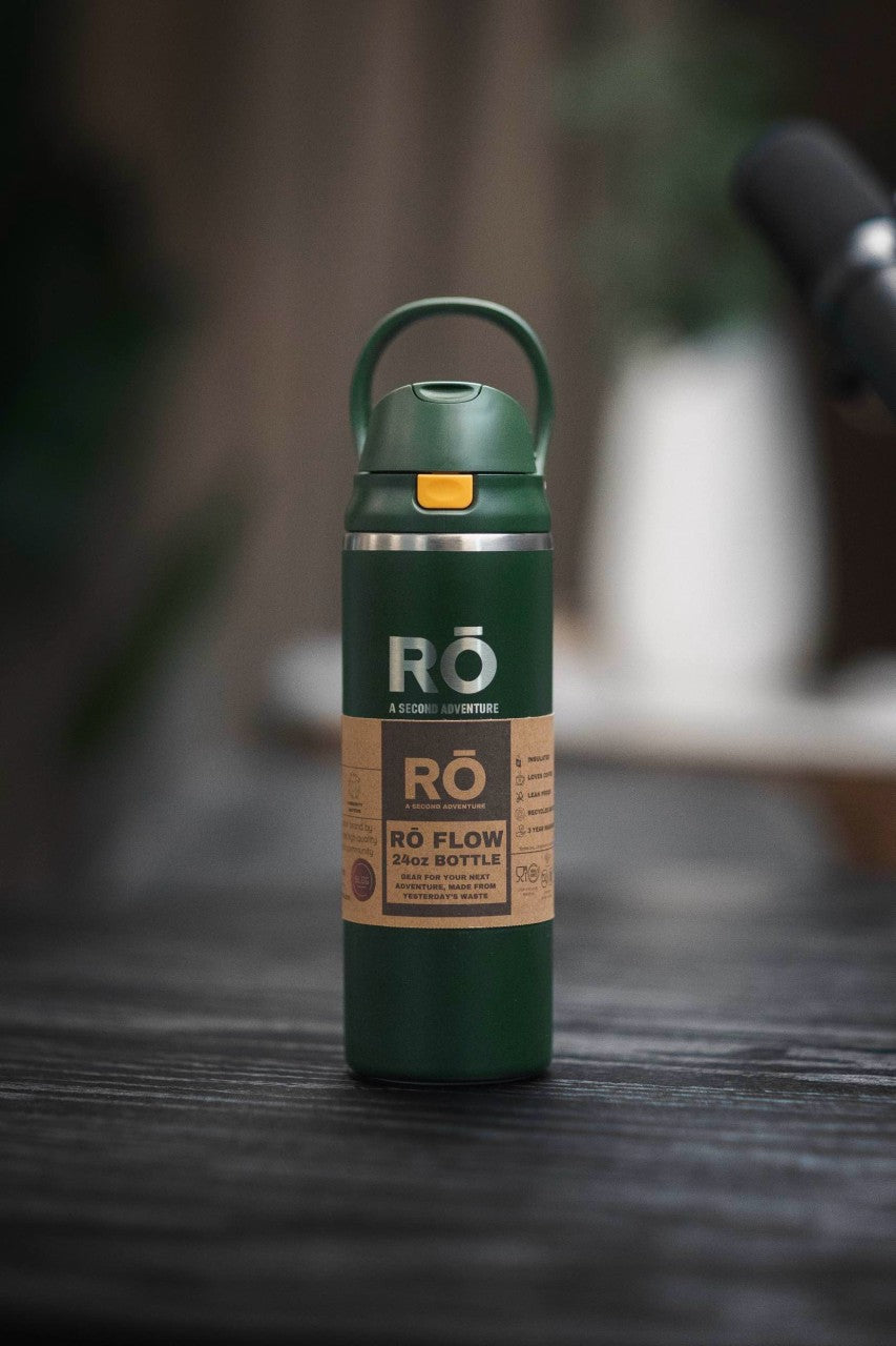 A dark green RO Flow 24oz bottle with a matching green loop handle and a tan label, standing on a dark wooden surface with a blurred background.