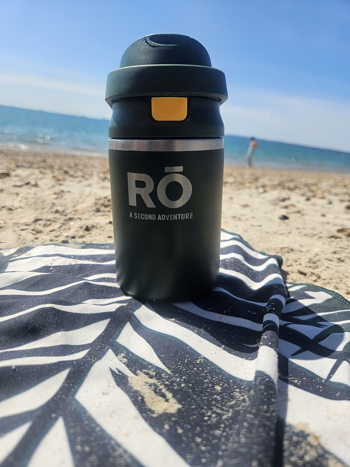 A matte black insulated travel tumbler with a flip-top lid stands on a black-and-white patterned beach towel on the sand at the beach, with the ocean and blue sky in the background. The tumbler has a silver band with the white text 'RO A SECOND ADVENTURE'.