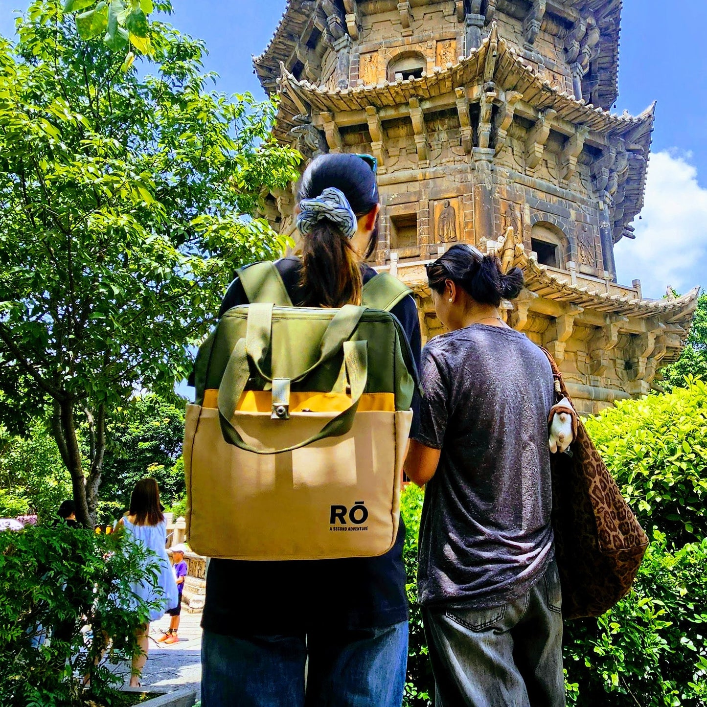 Two people viewed from behind standing outdoors in front of a large pagoda tower, with green trees and bushes around them and blue sky above. One person wears a tan backpack with a yellow-green top and blue jeans, and the other wears a dark shirt and dark pants.