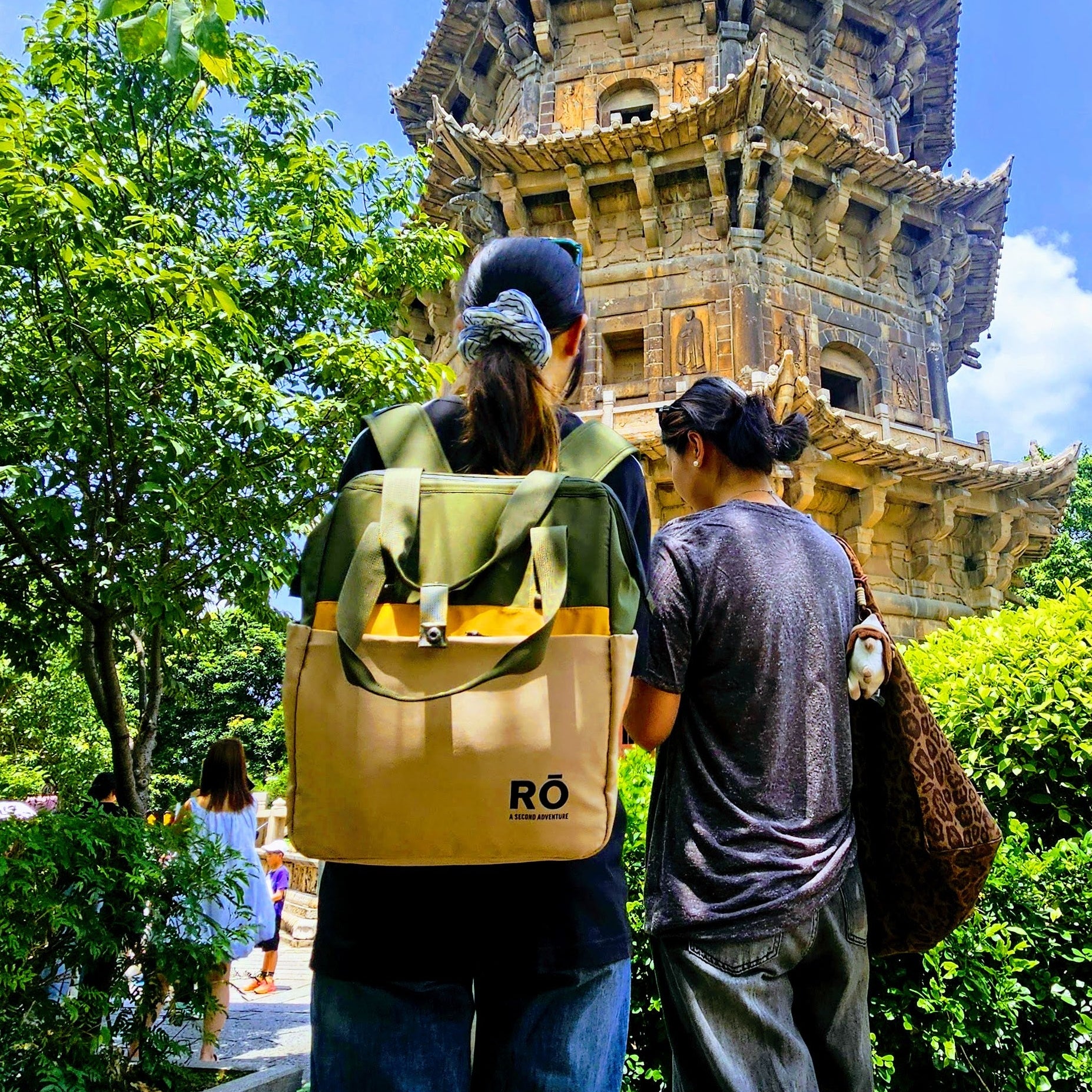 Two people viewed from behind standing outdoors in front of a large pagoda tower, with green trees and bushes around them and blue sky above. One person wears a tan backpack with a yellow-green top and blue jeans, and the other wears a dark shirt and dark pants.