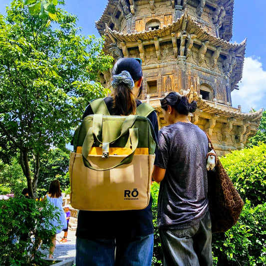 Two people viewed from behind standing outdoors in front of a large pagoda tower, with green trees and bushes around them and blue sky above. One person wears a tan backpack with a yellow-green top and blue jeans, and the other wears a dark shirt and dark pants.